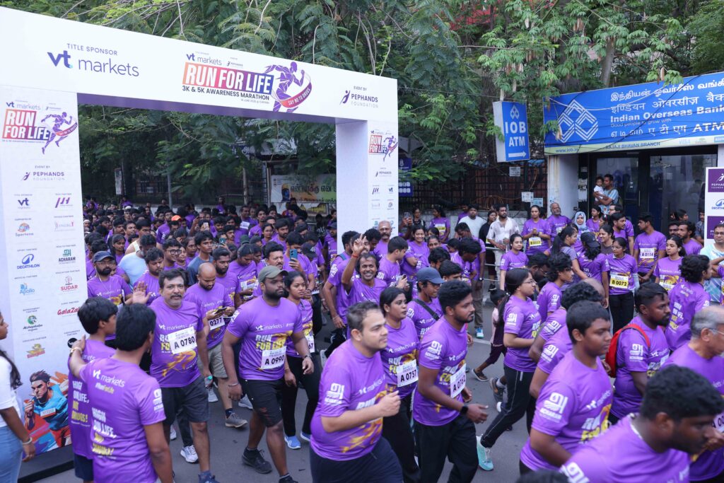 Crowd of runners in purple shirts gather at a marathon start under a sponsor banner reading Run for Life (3K & 5K). i