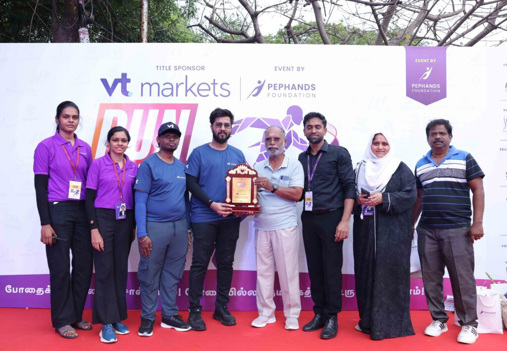 Group of people on a red stage posing with a trophy in front of a sponsorship banner at an event.